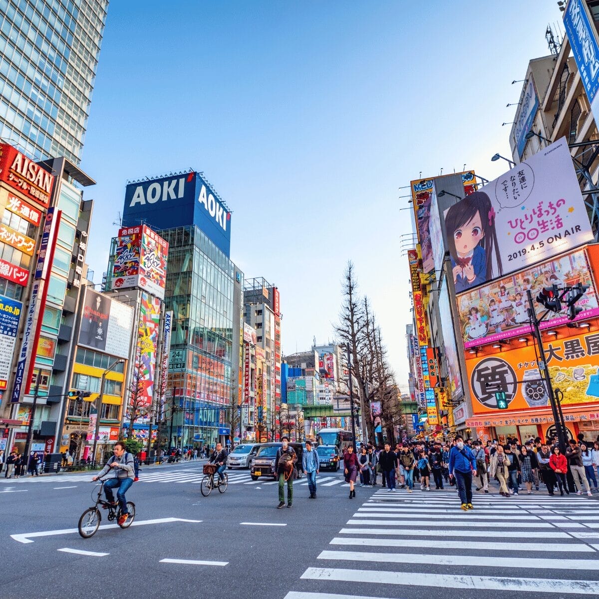 Cruce peatonal en Akihabara Tokyo con edificios AOKI y pantallas LED gigantes de anime, ciclistas y multitudes caminando en día soleado